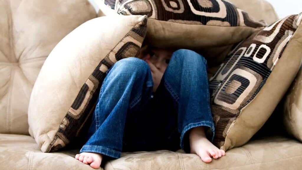 criança escondida entre almofadas sentada num sofá representando o medo