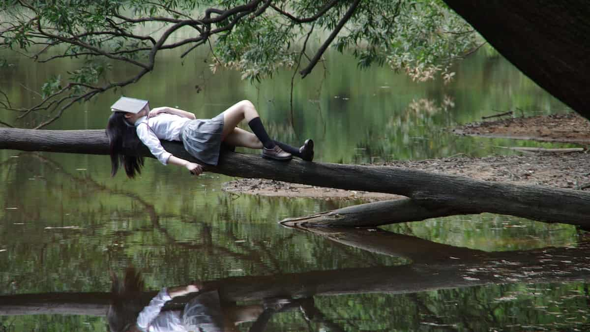 mulher deitada em cima de um tronco sob um rio, tendo um livro em cima da sua cabeça. Está dormindo